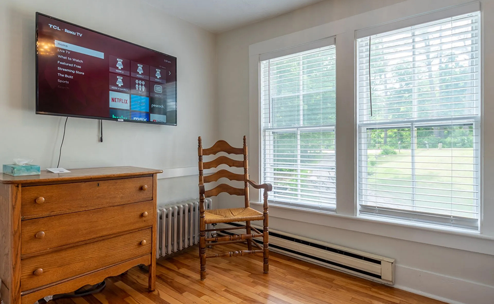 T V mounted on wall above an antique wooden dresser near a rocking chair and window